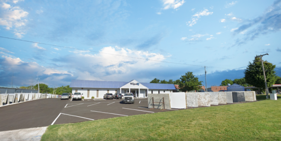 A modern, single-story commercial building with a white exterior and a blue metal roof sits behind a freshly paved parking lot. The sign above the entrance reads "Wilgus IQ." Large stone slabs are neatly arranged along the property, showcasing a variety of materials. To the right, a blue sign reads "SHOWROOM," indicating another business nearby. The sky is bright blue with scattered white clouds, and the surrounding area features well-manicured grass and trees. Several vehicles are parked in front, adding t
