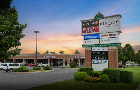 Aundria Plaza Monument Sign A detailed shot of the Aundria Plaza business directory sign in front of the shopping center at sunset. The sign lists businesses including The UPS Store, Rocco’s Pizza, Hinode, China King, Progressive, and more. Below the sign, manicured bushes and green grass surround the base. In the background, the shopping center features a brick exterior with arched walkways and parked cars. The sky displays a mix of warm sunset tones blending into cool blue.