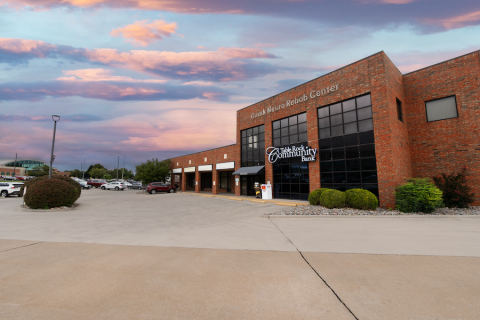 Bradford Centre Storefront A brick commercial building with large glass windows houses "Table Rock Community Bank" and "Ozark Neuro Rehab Center." The parking lot is mostly empty, with a few cars visible in the distance. Landscaping includes trimmed bushes and rock beds. The sky is a mix of pink and blue hues, indicating sunset.