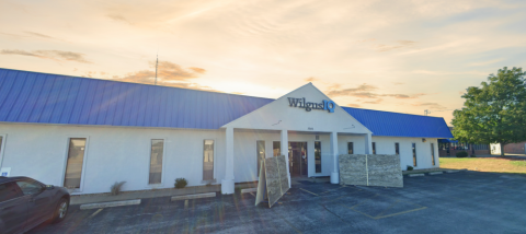 Casco-Scenic Storefront A single-story commercial building with a white exterior and a bright blue metal roof. The entrance features a triangular awning with the "Wilgus IQ" logo above the glass doors. The parking lot in front has a few spaces, with some construction materials leaning against the building. A large tree stands to the right, and the sky is a mix of golden hues, suggesting early morning or late afternoon.