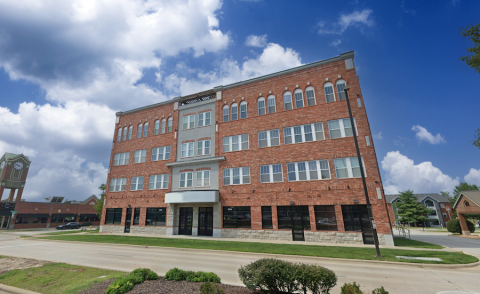 Chesterfield Lofts II A four-story red brick building with arched windows and a symmetrical design, situated along a landscaped street. The ground floor features large glass storefronts, ideal for commercial leasing. The sky is bright blue with scattered clouds, and a nearby shopping center is visible in the background.