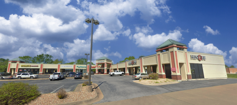 Crossroads Shopping Center - Exterior A slightly angled shot of Crossroads Shopping Center, showing various retail stores, including a restaurant with a visible "Hinode" sign. The architectural design features beige and red tones, decorative towers with green rooftops, and a parking lot with multiple vehicles. The sky is partly cloudy with blue patches visible.
